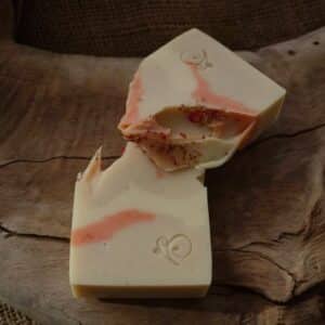 Two pieces of an artisanal cream-colored soap bar with coral pink swirls, resting on textured driftwood. Both pieces feature a small stamped logo, and the broken edge shows embedded dried botanicals.